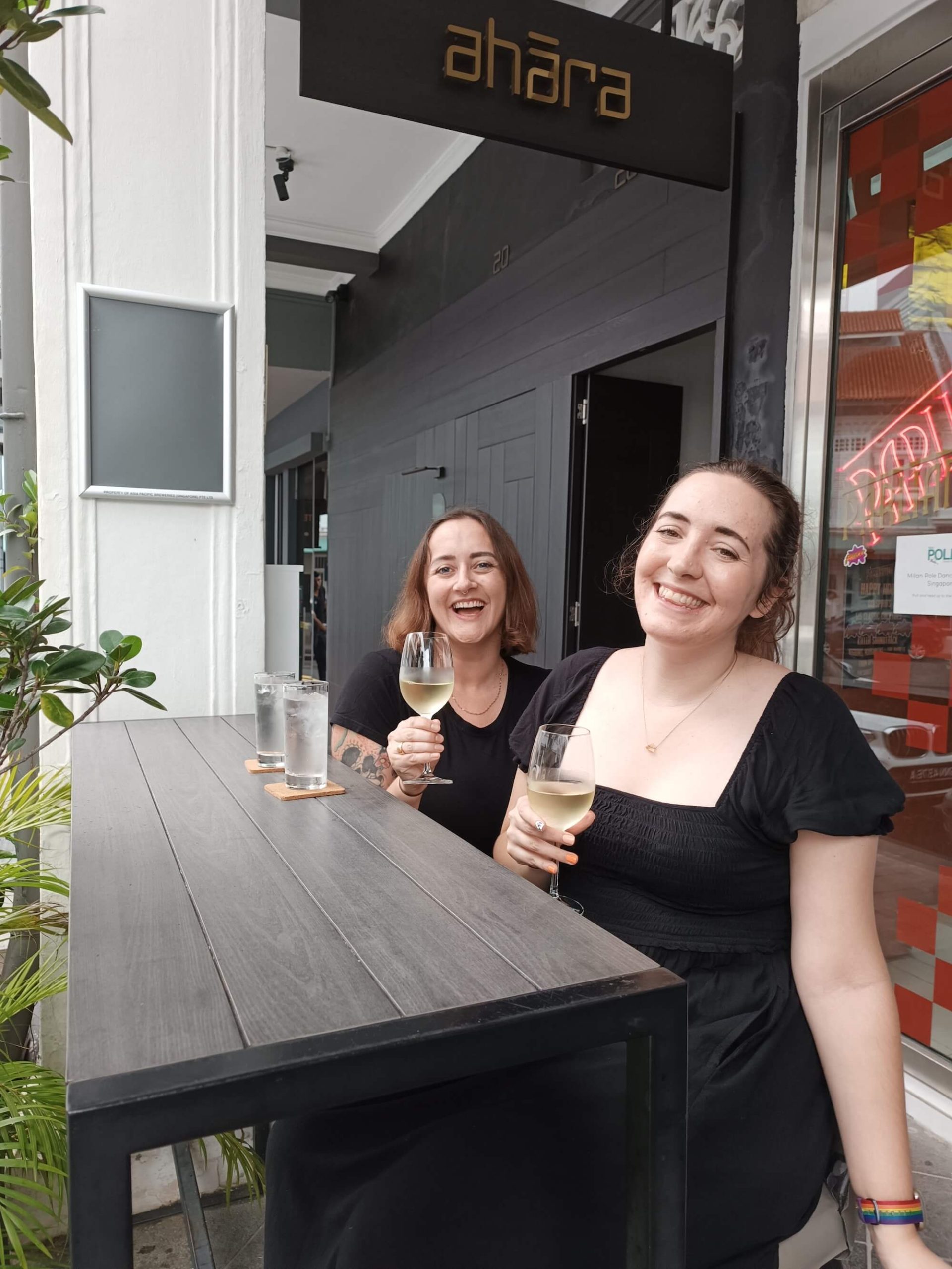 Holly & Sophie sit at an outdoor table both smiling and holding glasses of white wine