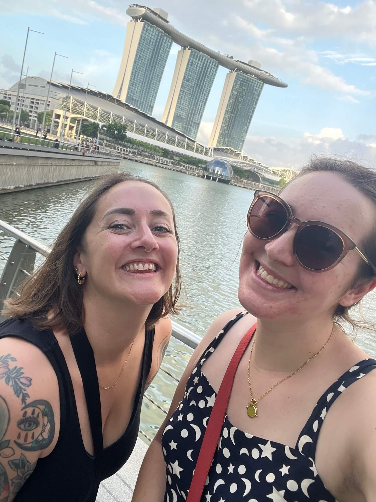 Sophie and Holly, two white women with short brown hair, are smiling and taking a selfie in front of the Marina Bay Sands hotel in Singapore.