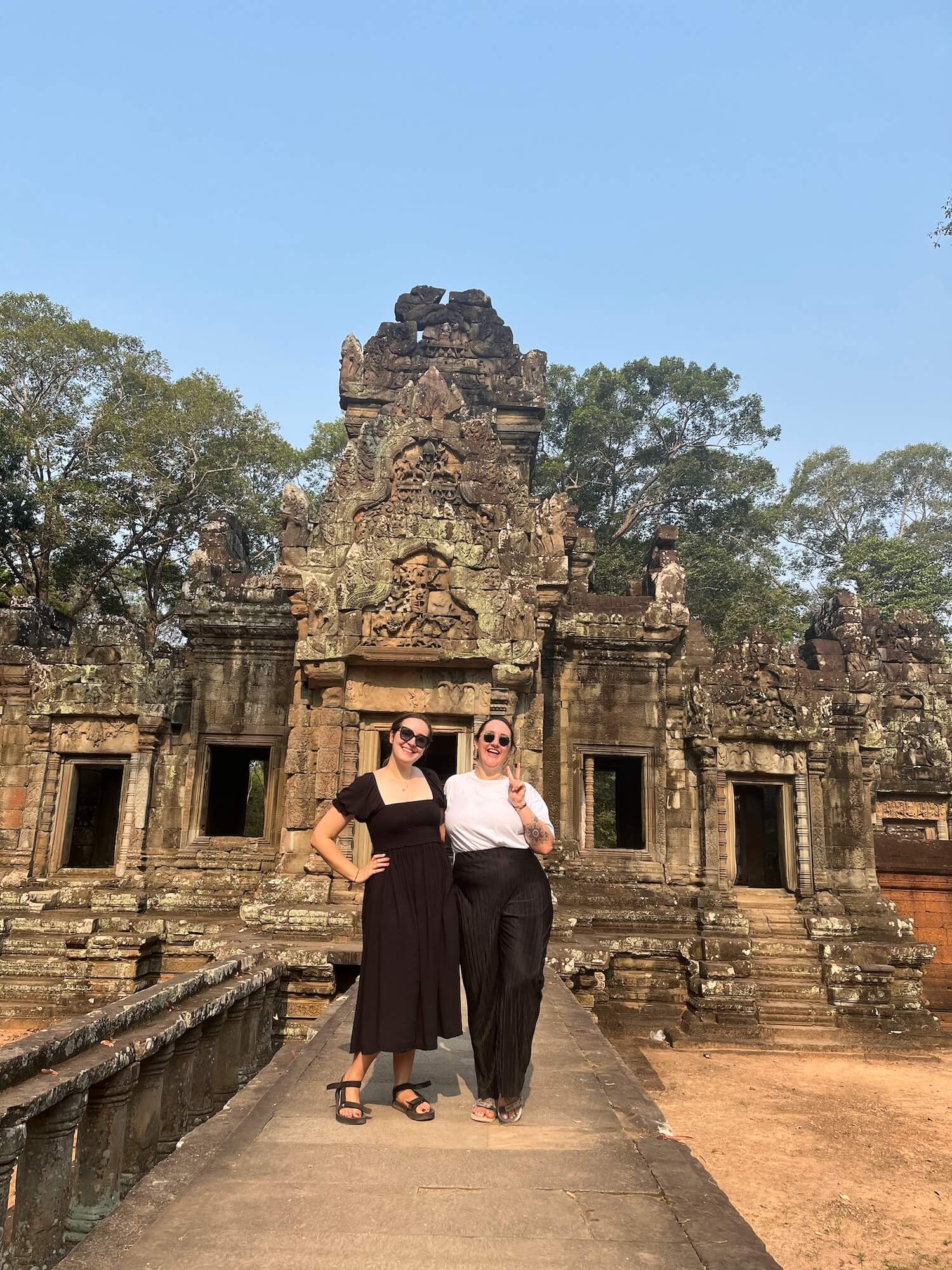 Sophie and Holly, two white women, are standing in front of an ancient temple at Angkor Wat in Cambodia. Holly is wearing a black dress and has a hand on her hip. Sophie is wearing a white t-shirt and black trousers and is doing a peace sign with her left hand.