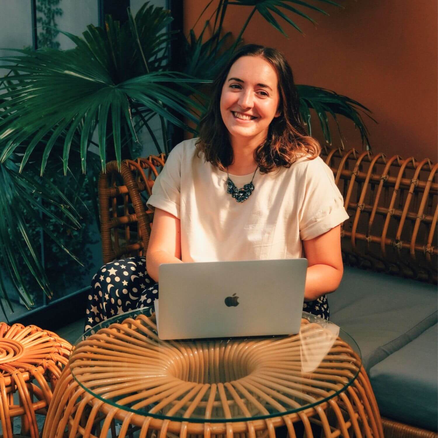 Holly, a woman with brown hair working on a laptop at a wicker table. She is smiling at the camera.