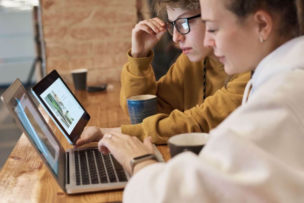 Two young people sit side-by-side at a light wooden high-top table, working collaboratively on open silver laptops. The person on the right, shown in close foreground, wears a white hoodie and has their hair tied back in a bun while typing on the keyboard. The person on the left wears black-framed glasses and a mustard yellow sweatshirt, leaning in closely to point at a website layout on the screen. Two coffee mugs sit on the table, with a warm, softly blurred brick wall in the background.