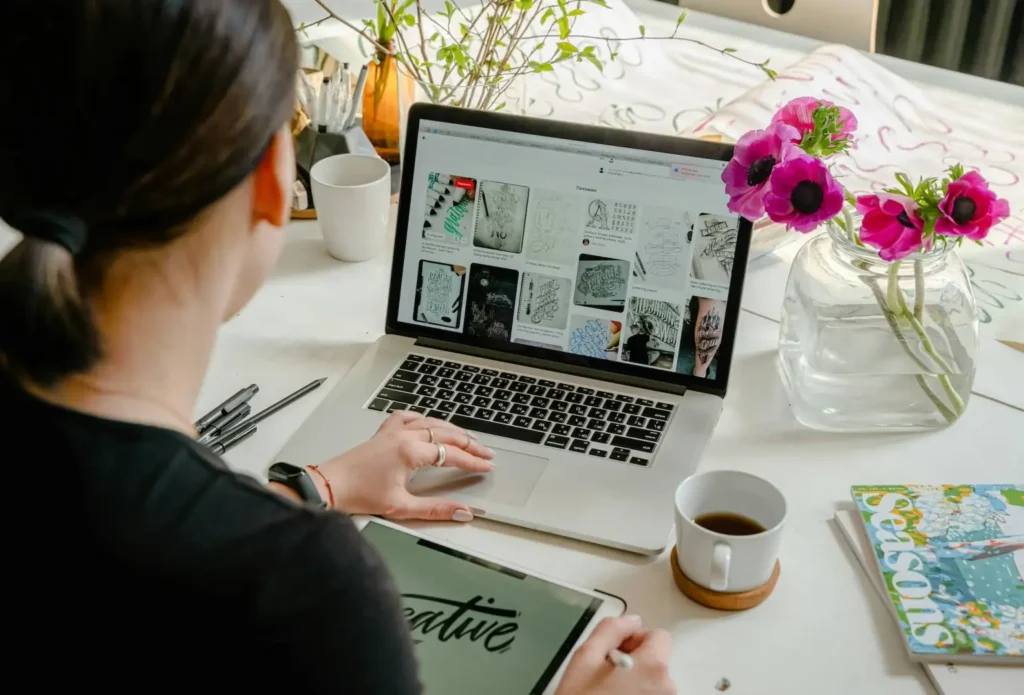 A designer sitting at a desk with flowers and coffee, browsing design references on her laptop while referencing a web design glossary.